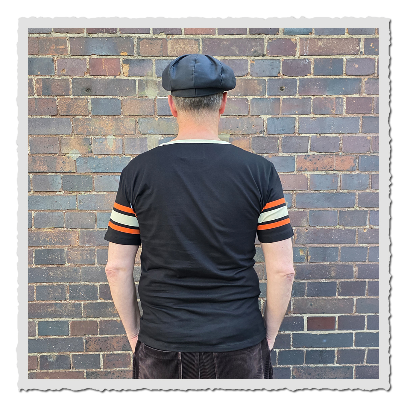 Rückenansicht einer Person mit schwarzem Shirt und Mütze vor rustikaler Ziegelwand.